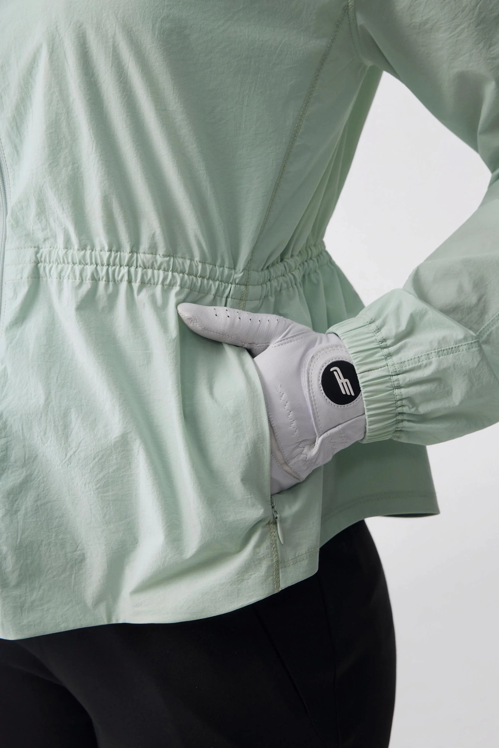 Close-up of a golfer placing a gloved hand in the zip pocket of a sage mist lightweight golf jacket
