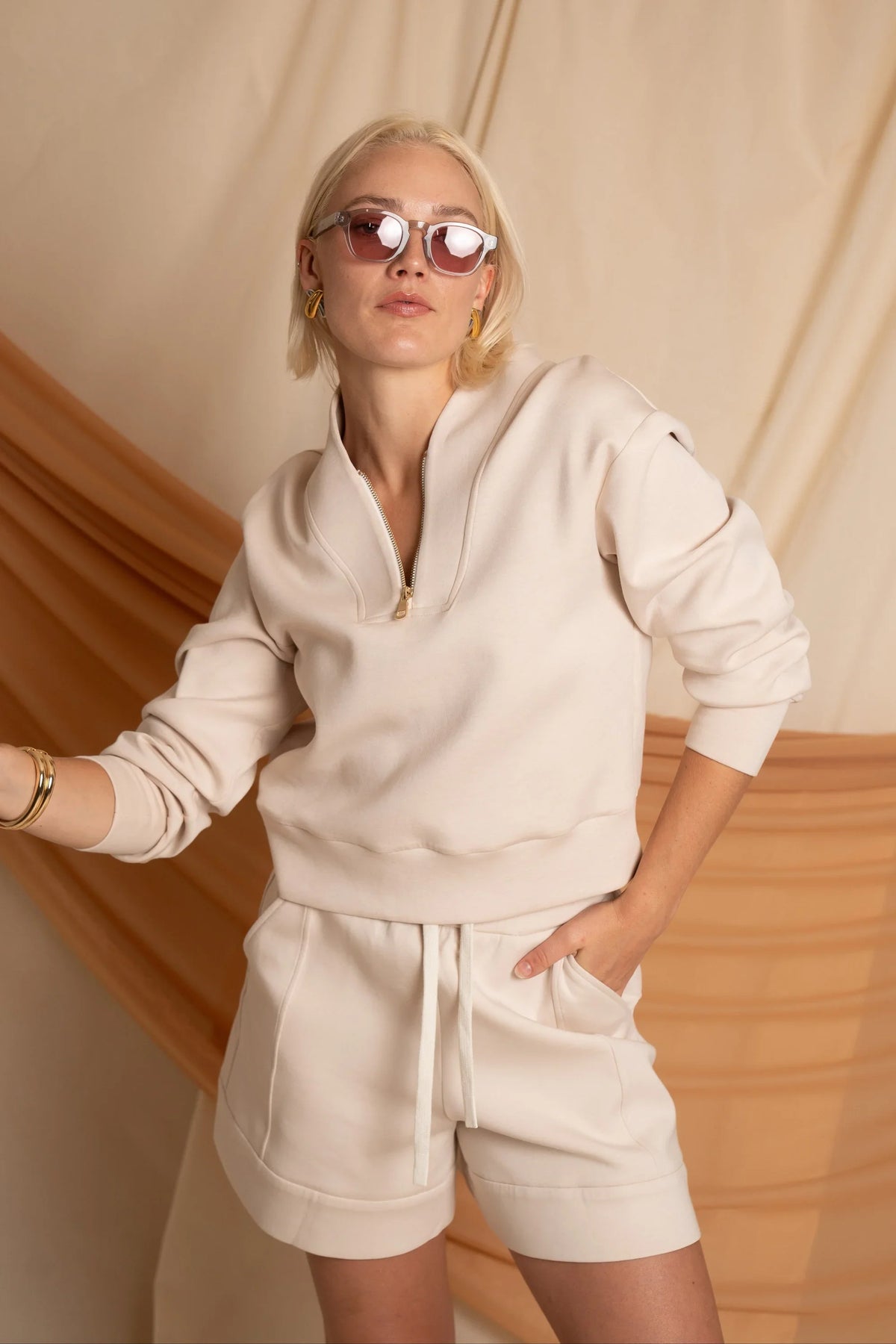 Woman wearing a cream half-zip golf pullover with matching shorts and sunglasses in an outdoor setting