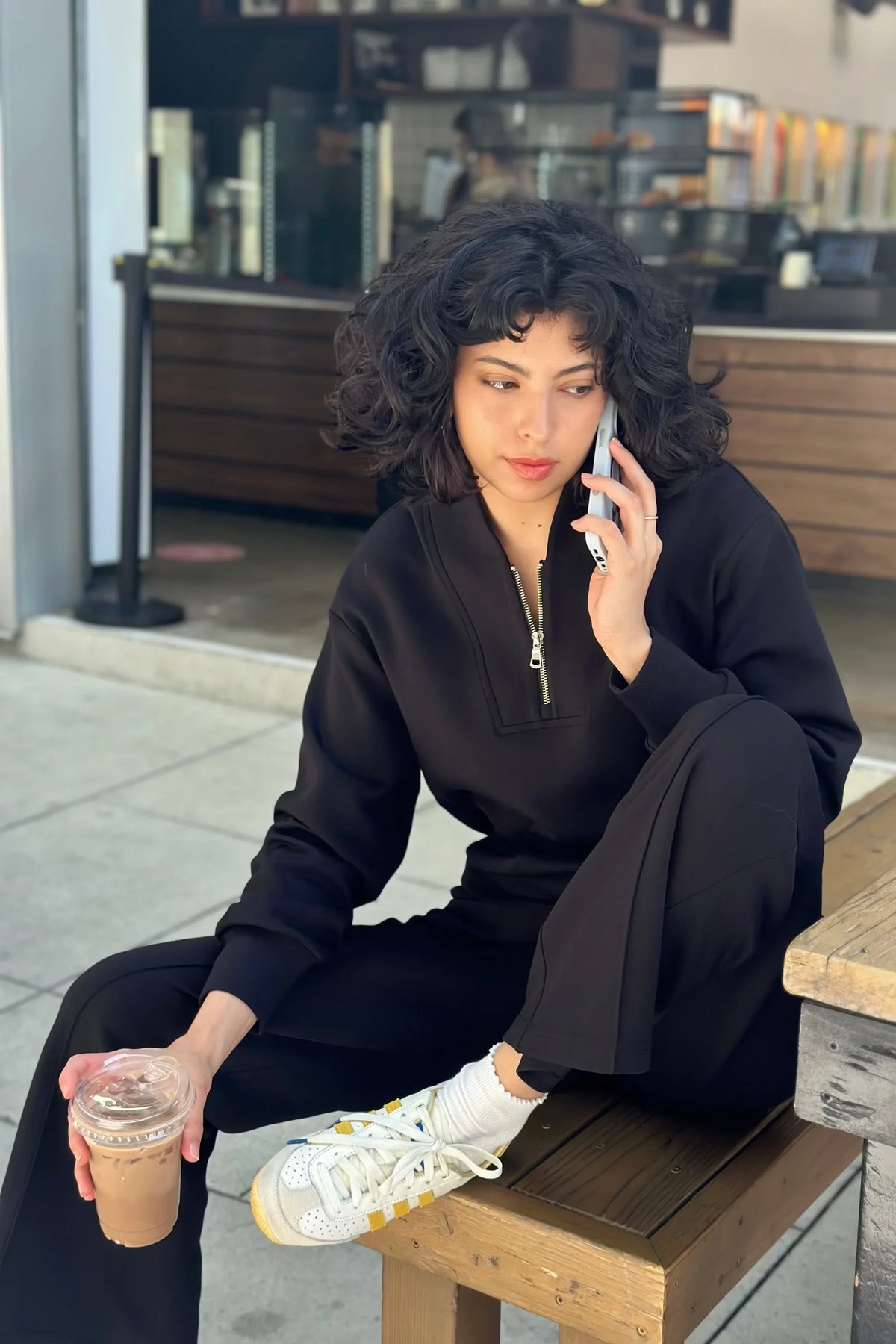 Woman wearing a black half-zip golf pullover with matching pants and sneakers while enjoying coffee outdoors