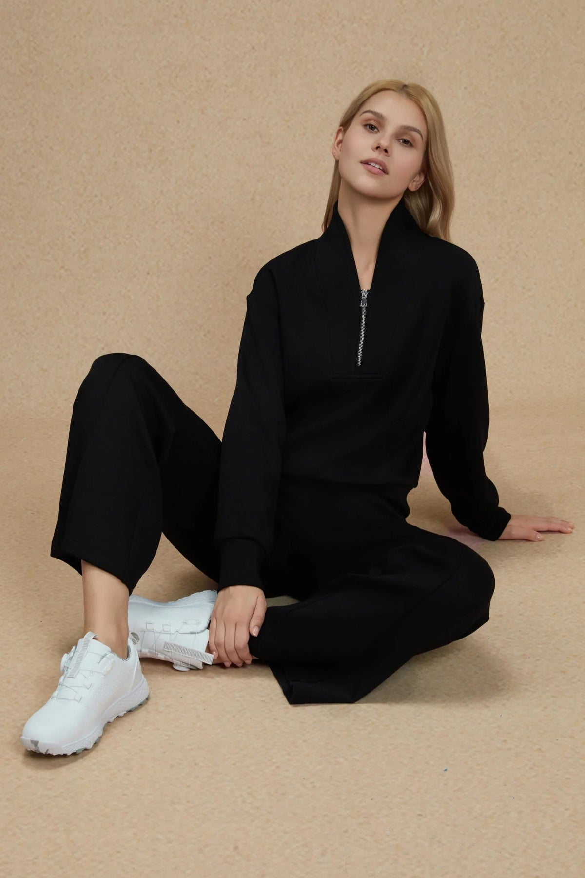 Woman seated in a black half-zip golf pullover and matching straight-leg pants with white sneakers