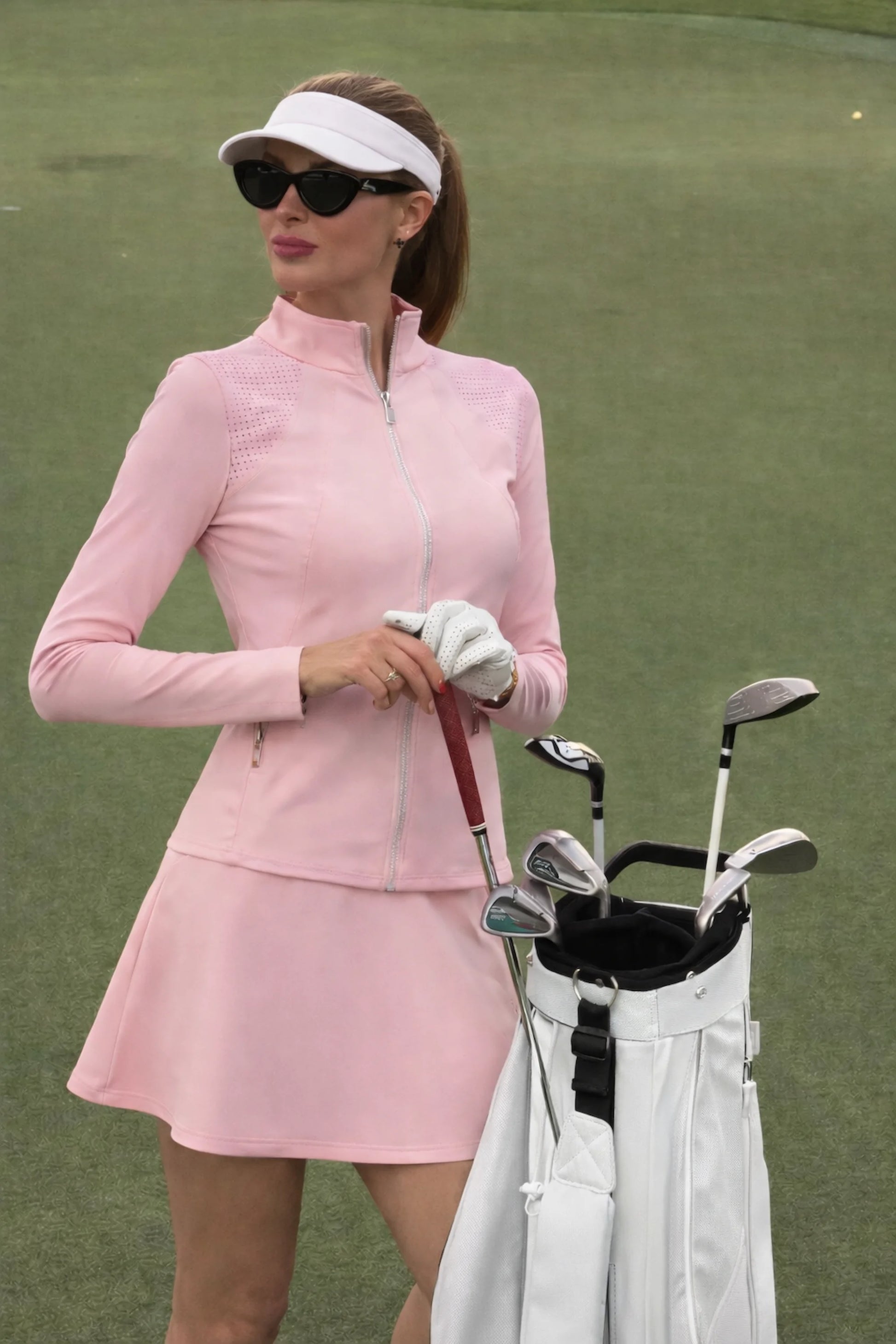 Model wearing women’s peplum zip golf jacket with mesh panels in pink on the golf course beside a golf bag