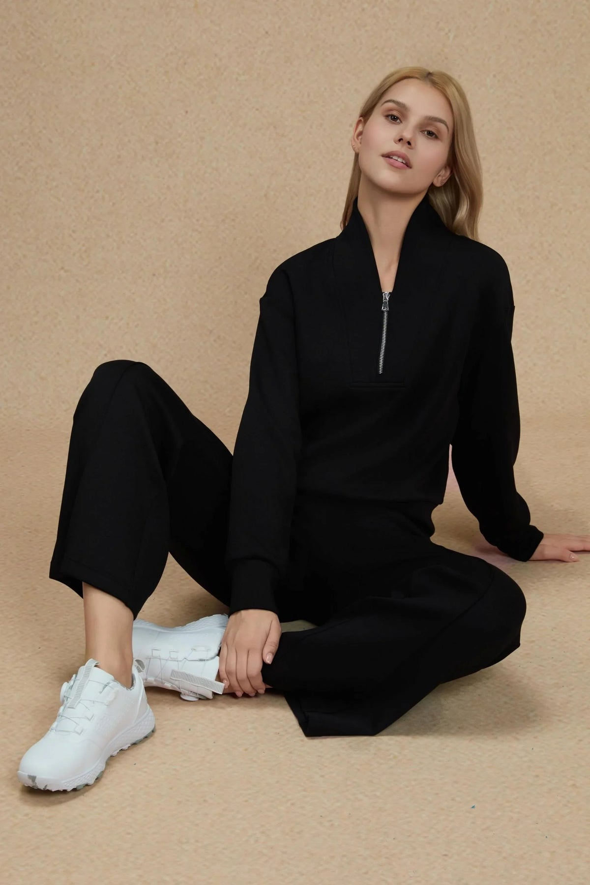 Woman seated in a black coordinated outfit featuring relaxed straight-leg pants and white sneakers