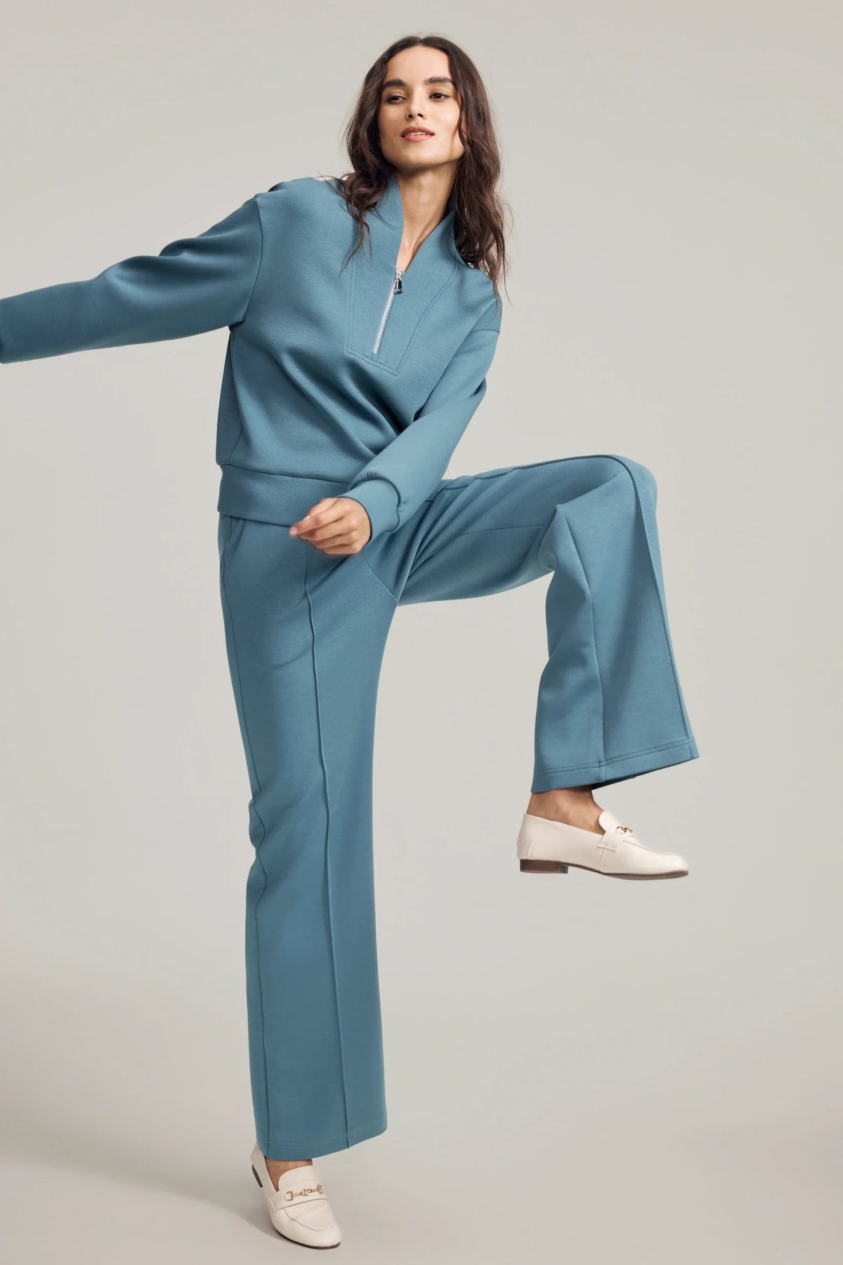 Woman wearing Slate Blue straight-leg pants with a matching pullover in a playful raised-leg pose
